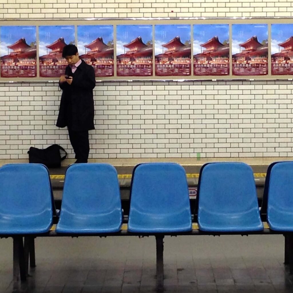 Japanese salaryman waiting in train station in black coat, using his phone. The seating area features bright blue chairs, and colorful advertisements enhance the modern, busy transportation hub ambiance.