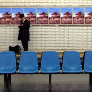 Japanese salaryman waiting in train station in black coat, using his phone. The seating area features bright blue chairs, and colorful advertisements enhance the modern, busy transportation hub ambiance.