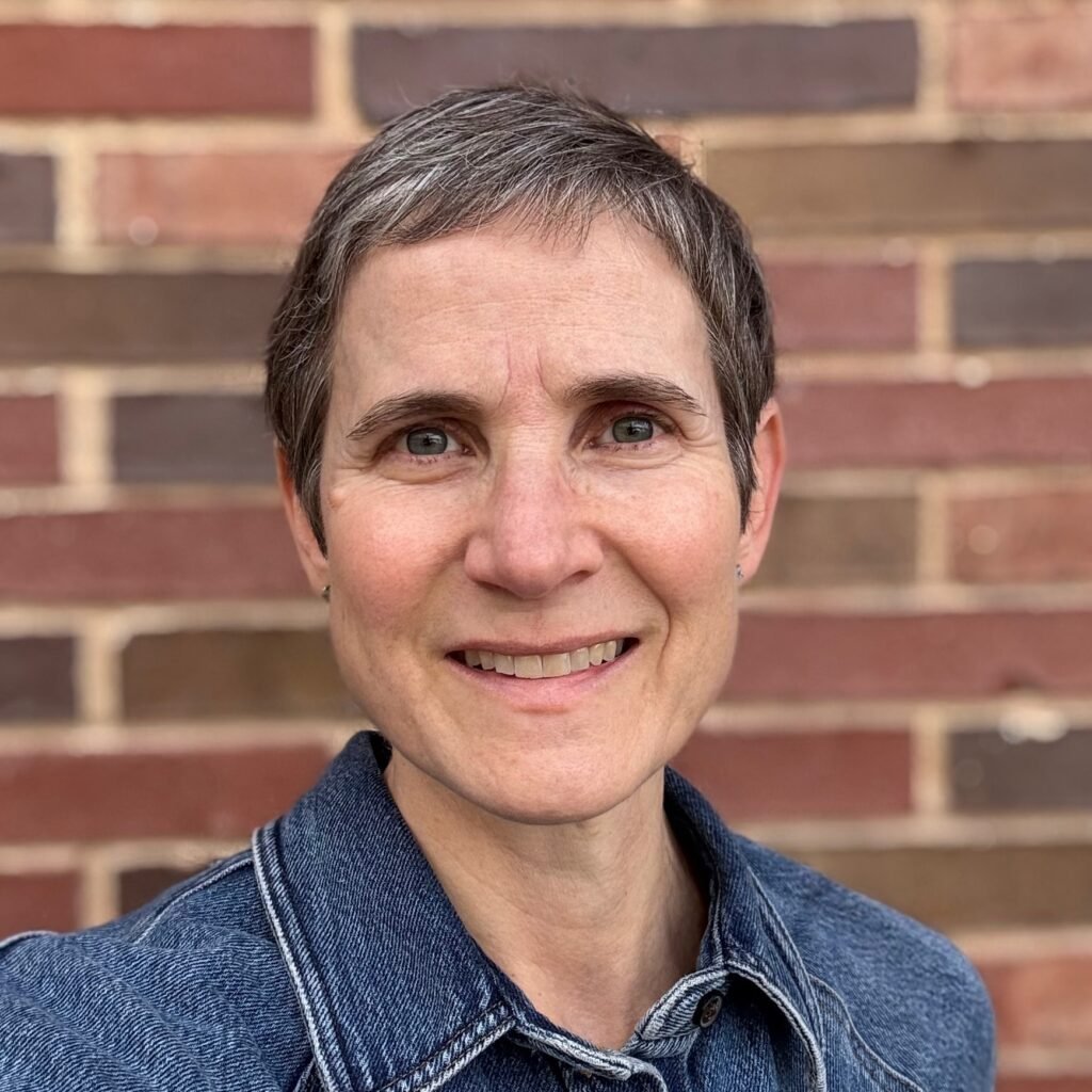Certified Travel Coach Linda from Create.Travel.Evolve. wearing a denim jacket, standing outdoors in front of a brick wall, conveying a sense of joy, confidence, and vibrant lifestyle.