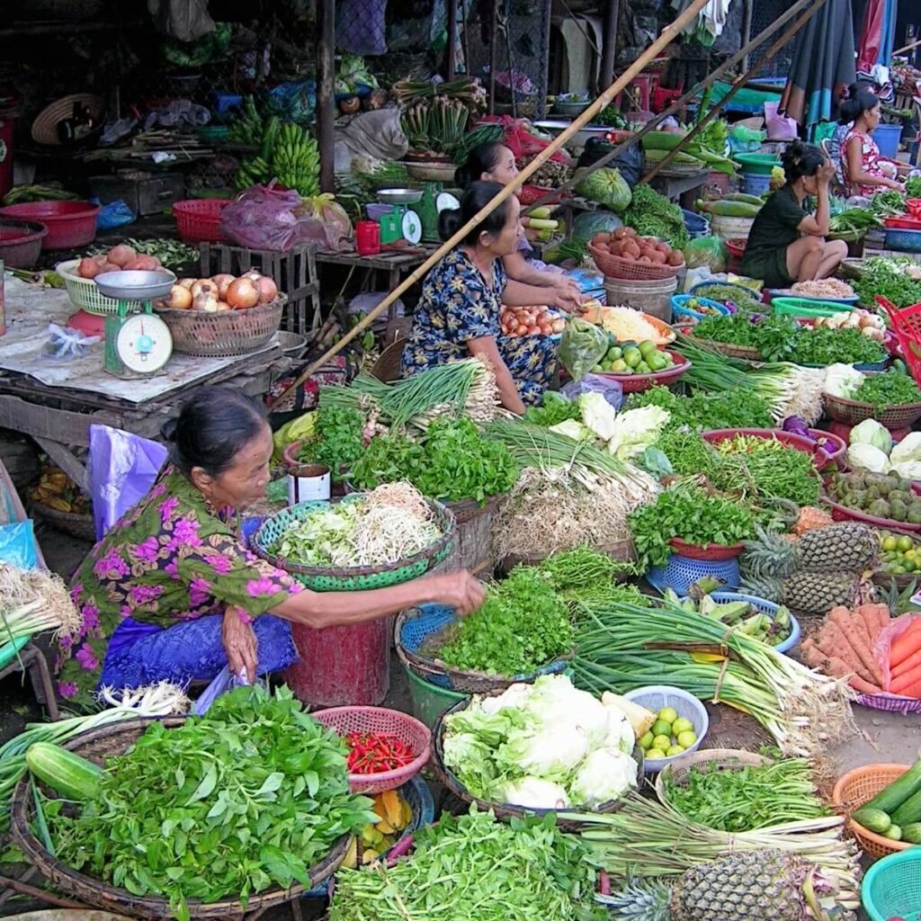 A colorful open-air market in Vietnam with local vendors selling fresh vegetables and fruits, including lettuce, onions, and herbs, in a lively, bustling environment ideal for travelers seeking authentic cultural experiences.
