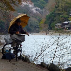 A man sits on his bicycle by a river, painting while holding an umbrella on a rainy day amid scenic natural surroundings in Arashiyama Japan.