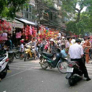 Vibrant Hanoi street market scene with shoppers and motorbikes amidst colorful vendors and decorations in a bustling environment.